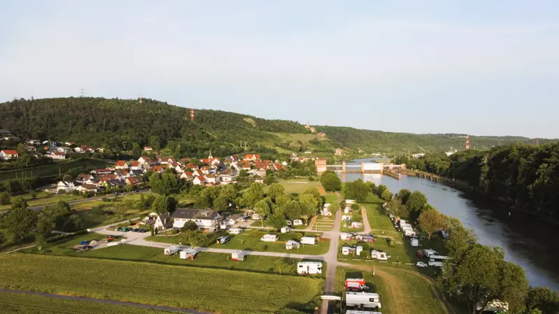 Luftaufnahme des Campingplatzes am Neckar mit dem Ort und bewaldeten Hängen im Hintergrund