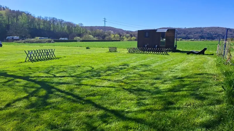 Gemähte Wiese mit niedrigen Holzzäunen, im Hintergrund eine dunkle Holzhütte und bewaldete Hügel