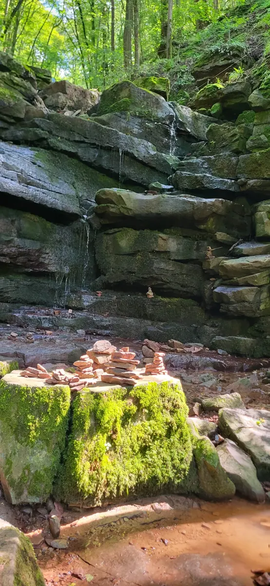 Geschichtete, moosbewachsene Felsstufen mit dünnen Wasserläufen in der Margaretenschlucht, im Vordergrund kleine Steintürmchen auf einem bemoosten Felsblock